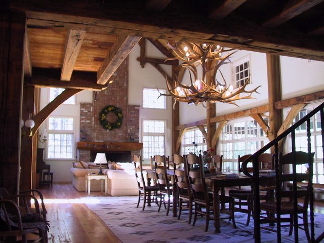 Spacious dining room with wooden beams and antler chandelier, overlooking living room with fireplace and windows.