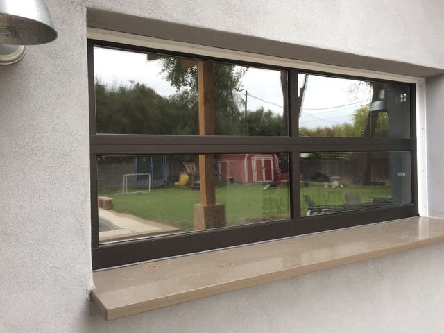 Brown-framed window with reflective glass, set in a white wall with a stone ledge, reflecting a backyard view.
