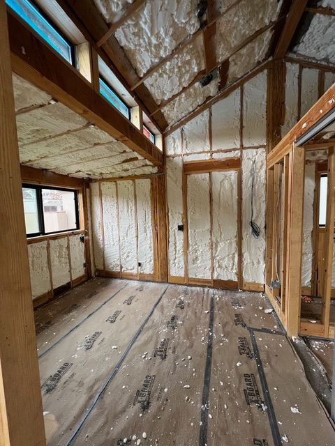 Interior of a building under construction, walls and ceiling insulated with foam, wood framing visible, covered floor.