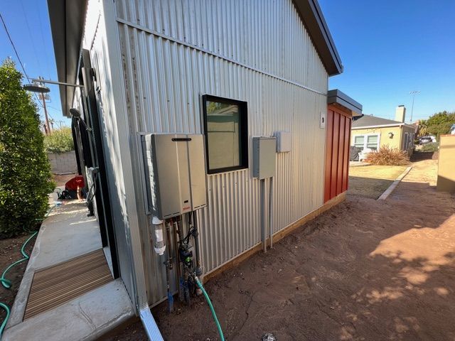 Exterior of a modern building with corrugated metal siding, water heater, red door, and a clear blue sky.