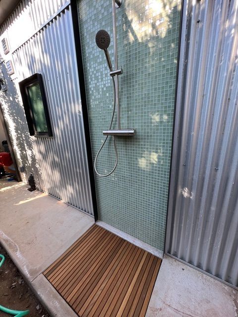 Outdoor shower with teal tile backing, corrugated metal walls, and wooden floor mat.