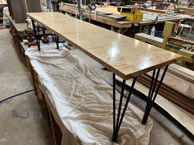 Long, finished wooden table with black hairpin legs in a workshop setting.