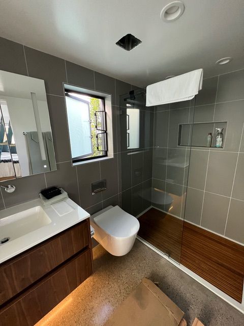 Modern bathroom with grey tile, wooden vanity, and shower. White toilet, open window.