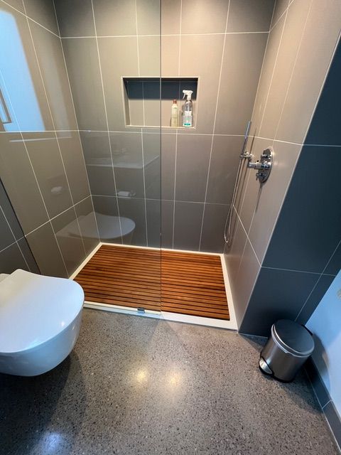 Modern bathroom with gray tiled walls, teak shower mat, and a white toilet.