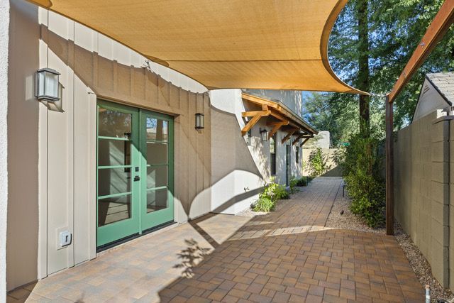 Beige building with green double doors, brick walkway, and a tan sun shade.