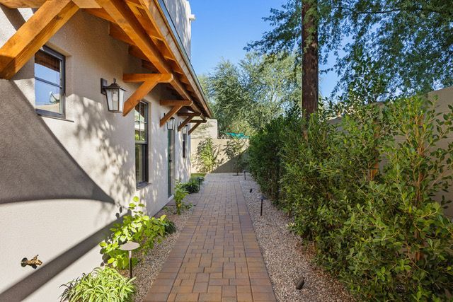 Brick pathway alongside a beige building with wooden awning, bordered by shrubs and gravel.