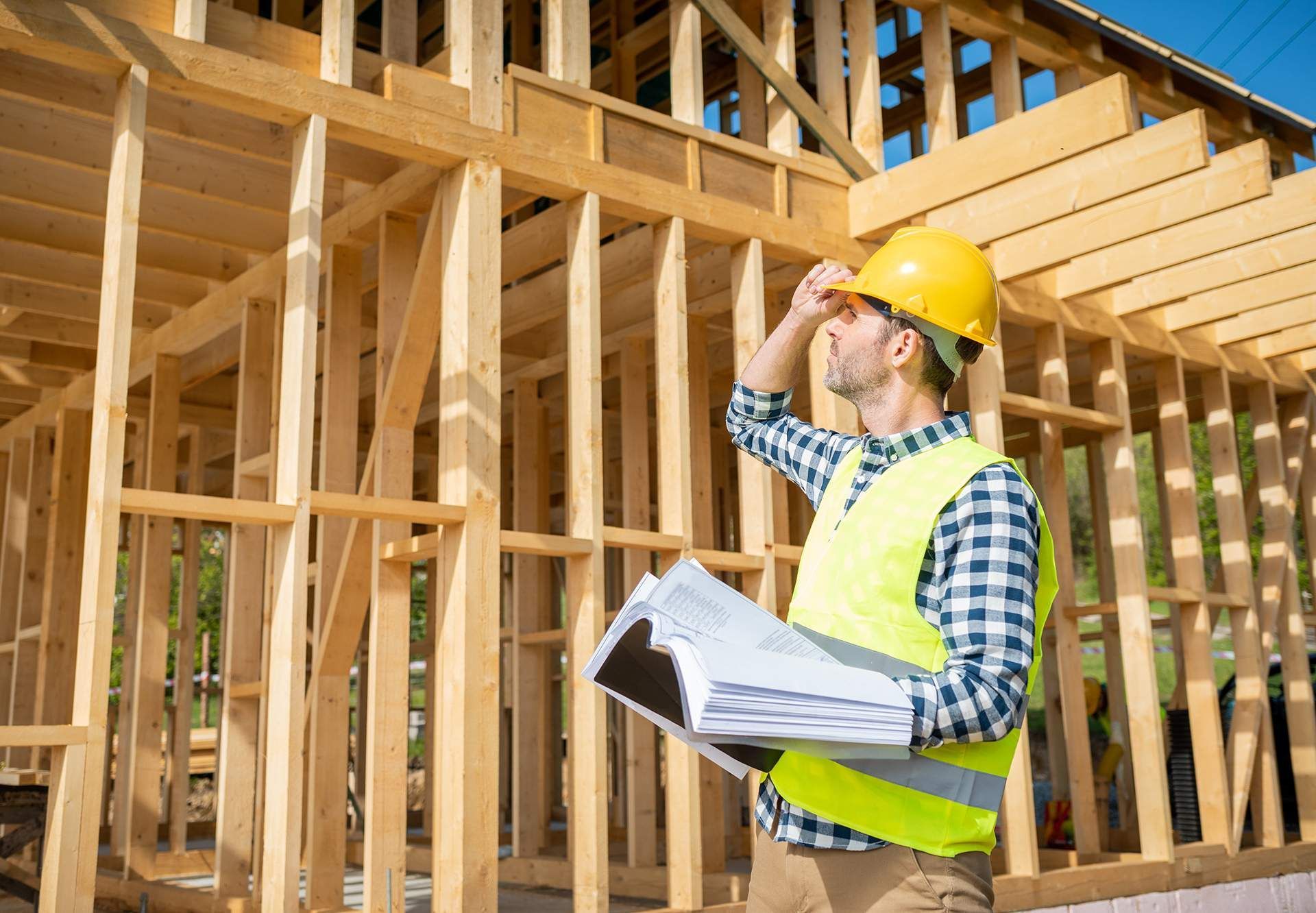 A construction worker is standing in front of a wooden house under construction.