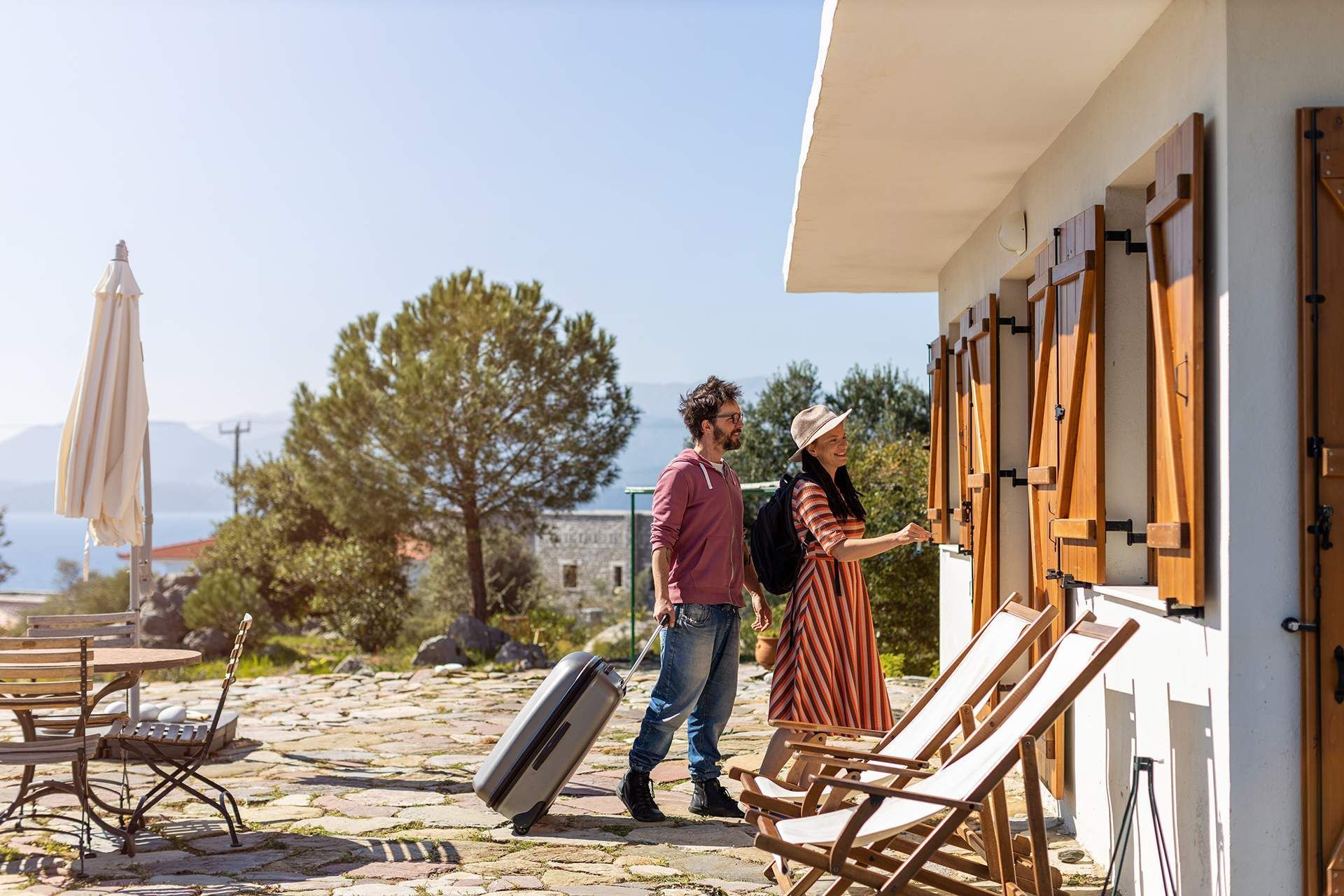 A man and a woman are standing in front of a house with a suitcase.