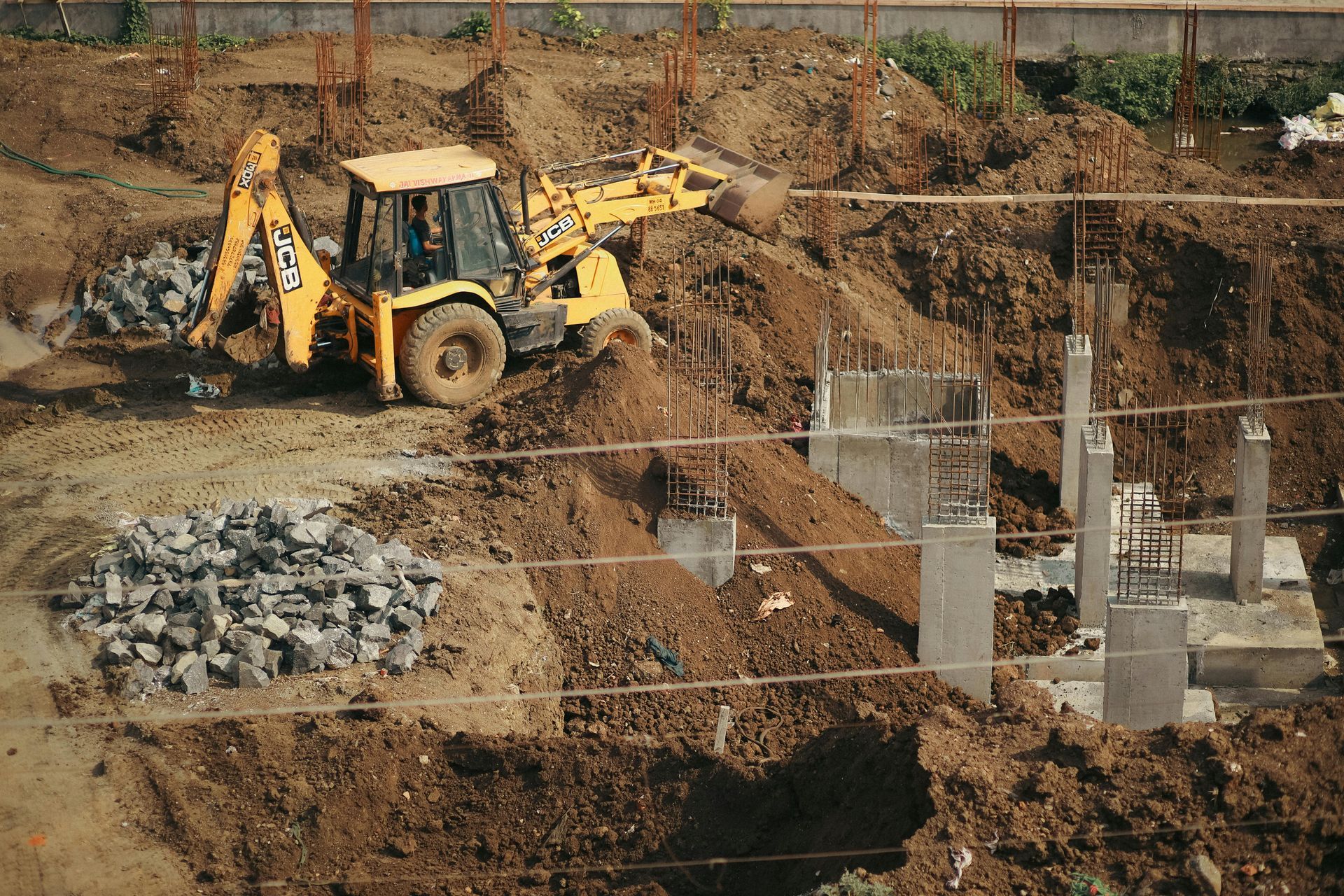 Yellow backhoe moving dirt on a construction site with concrete pillars and a pile of stones.