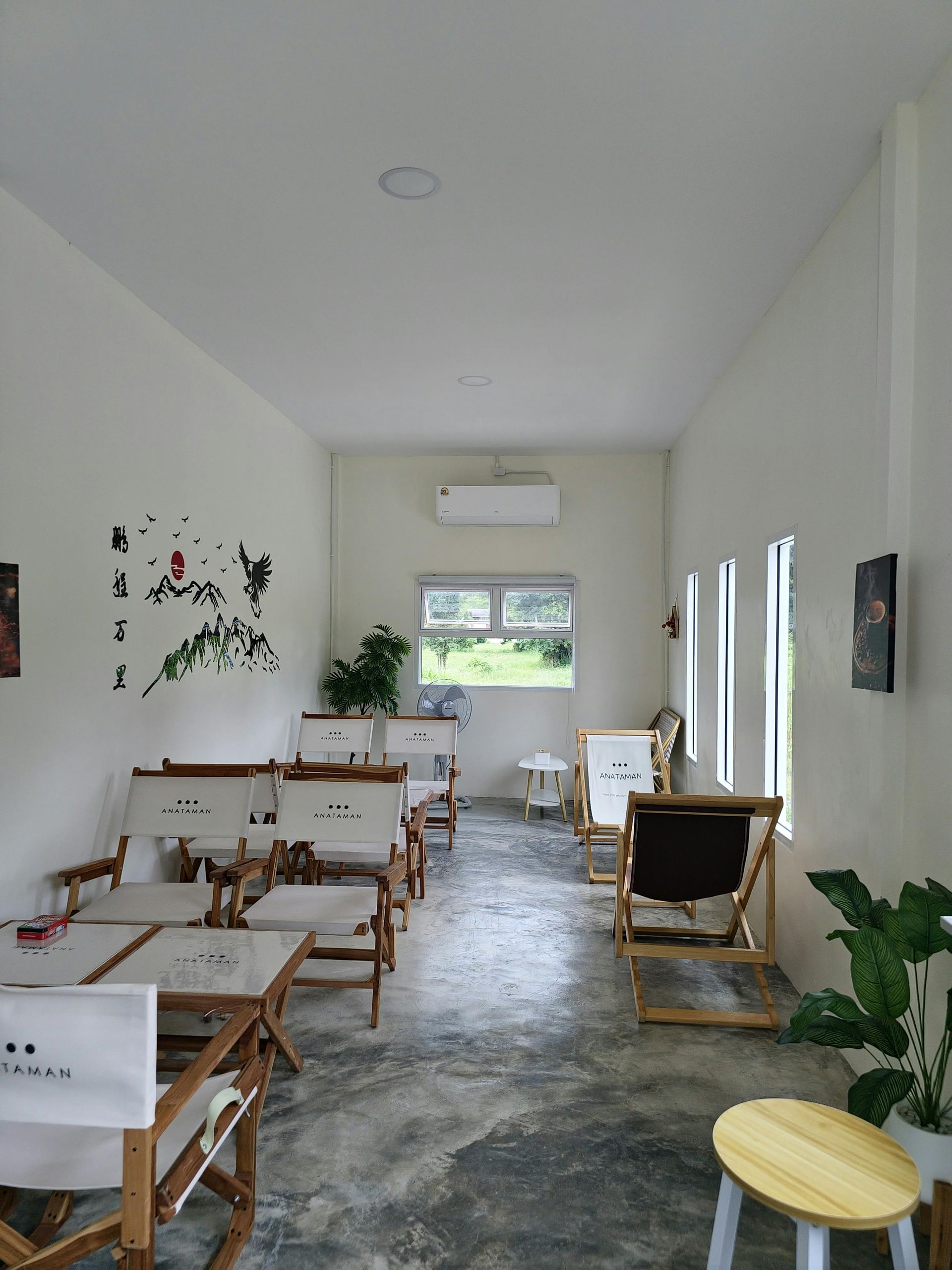 Cafe interior: white walls, wooden chairs, concrete floor, small windows, potted plants.