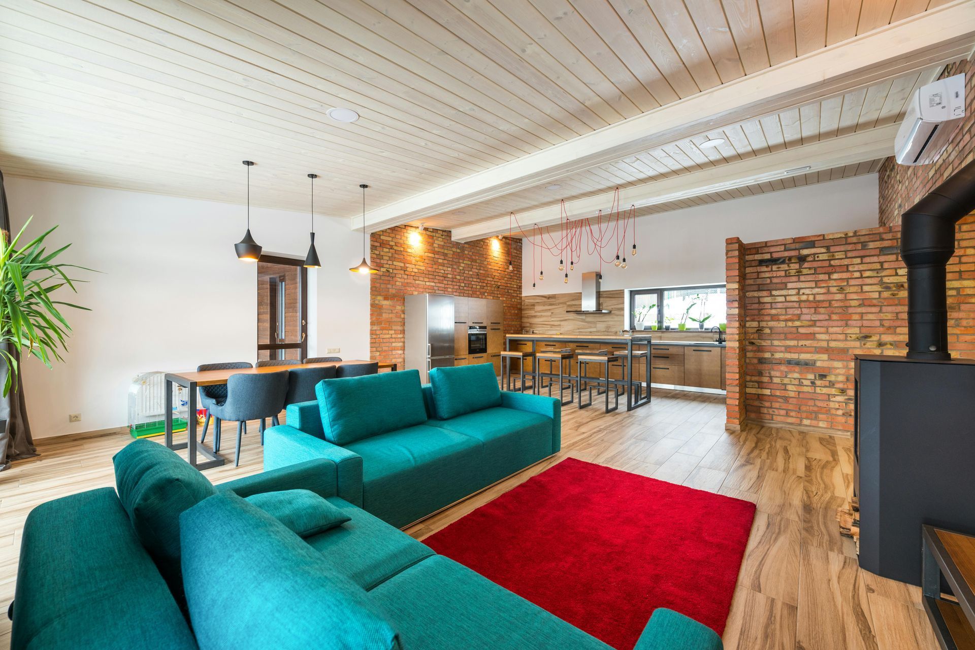 Spacious open-plan living room with teal sofa, red rug, and exposed brick walls.
