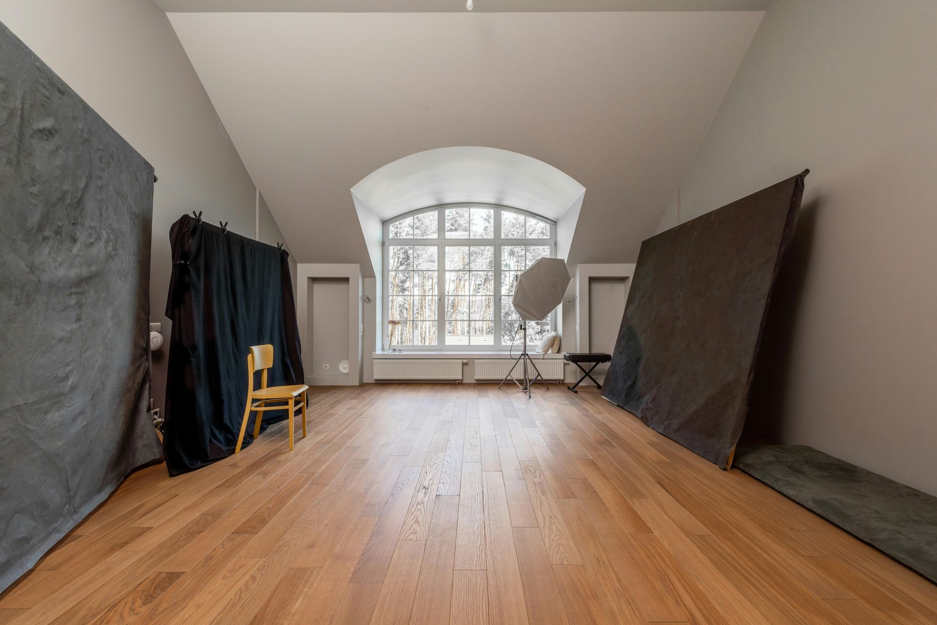 Empty photo studio with arched window, wooden floor, backdrops, and lighting equipment.