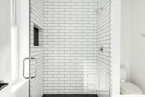 Modern white-tiled walk-in shower with glass door and black floor tile in a bright bathroom