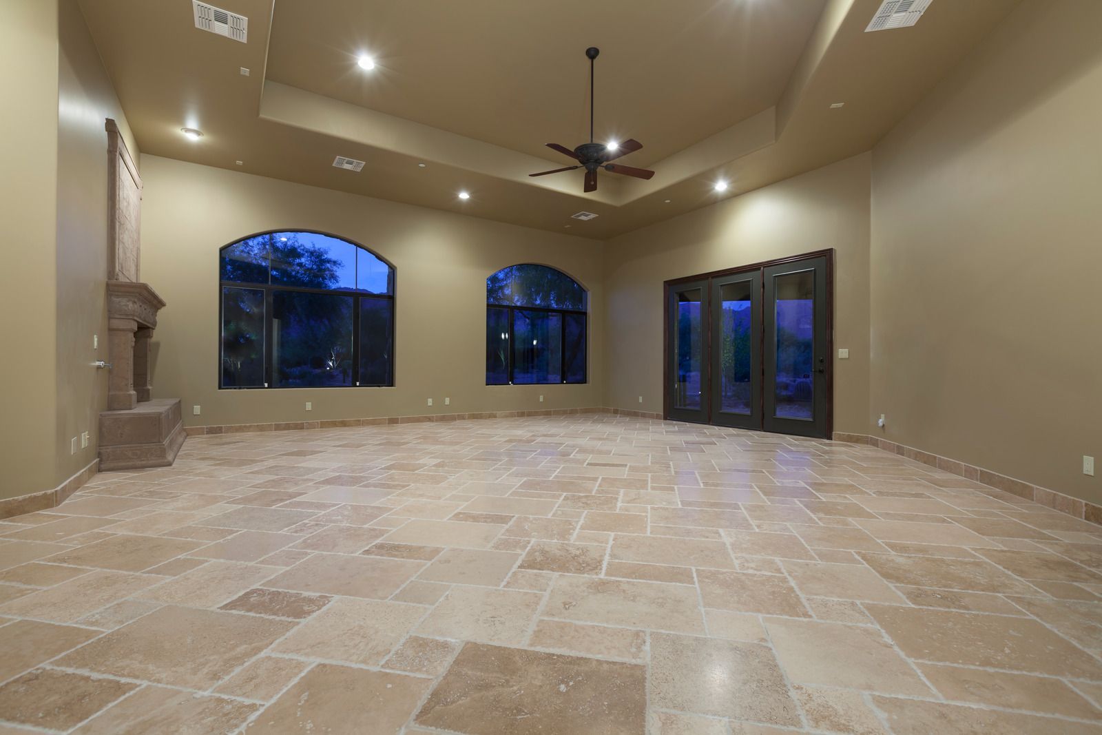 Empty beige living room with tiled floor, recessed lights, ceiling fan, and large arched windows and glass doors