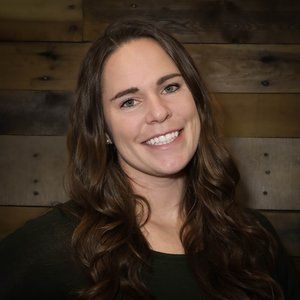 A woman with long brown hair is smiling in front of a wooden wall.