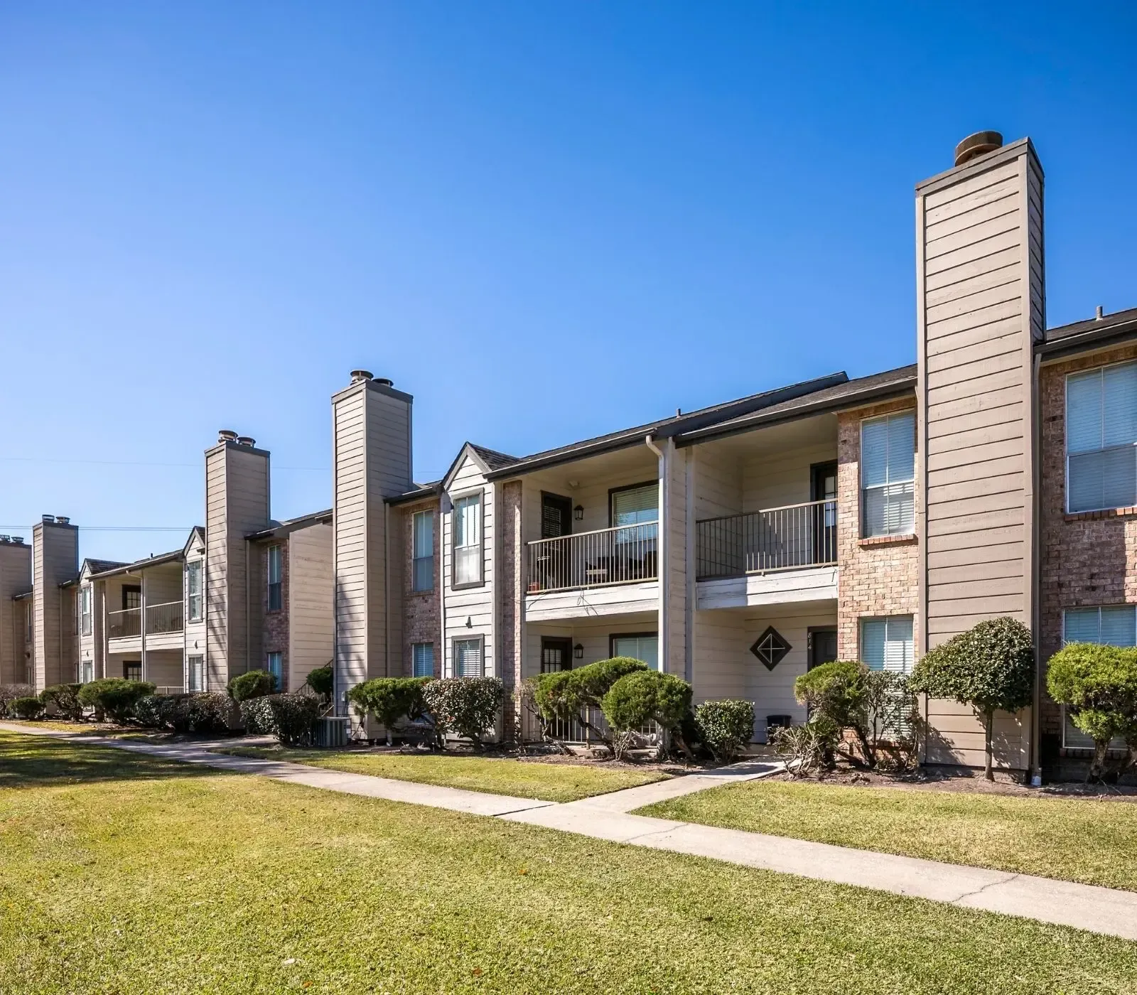 Apartment complex with brick and tan siding, chimneys, balconies, green lawn, and blue sky.