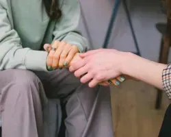 Two people hold hands in a supportive gesture, one seated in gray pants and the other in a plaid shirt.