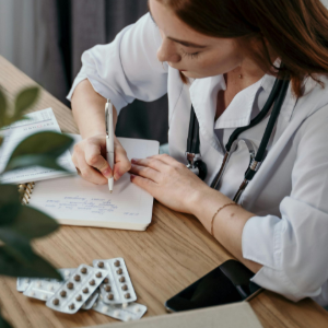 Healthcare professional writing notes at a desk with a stethoscope, pill blister packs, and a phone nearby