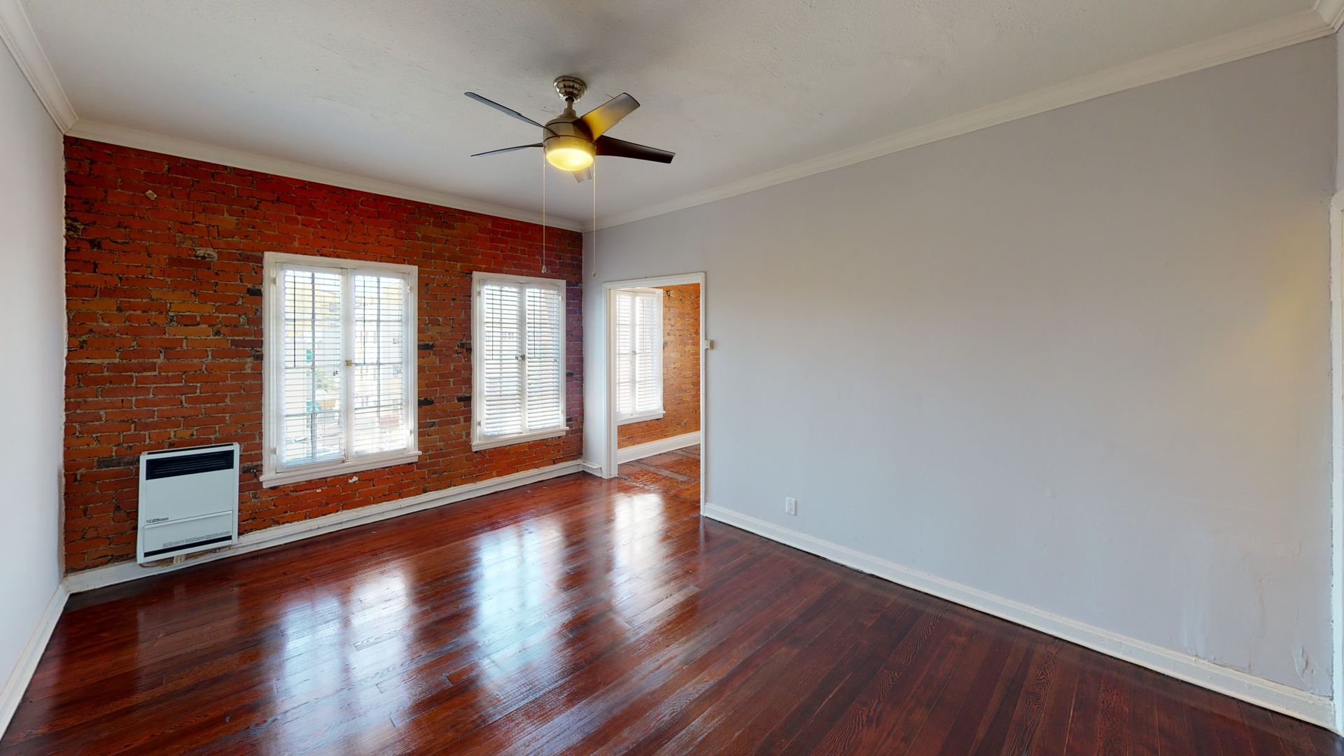 Interior view of a room with brick wall, windows, wooden floor, and a ceiling fan.