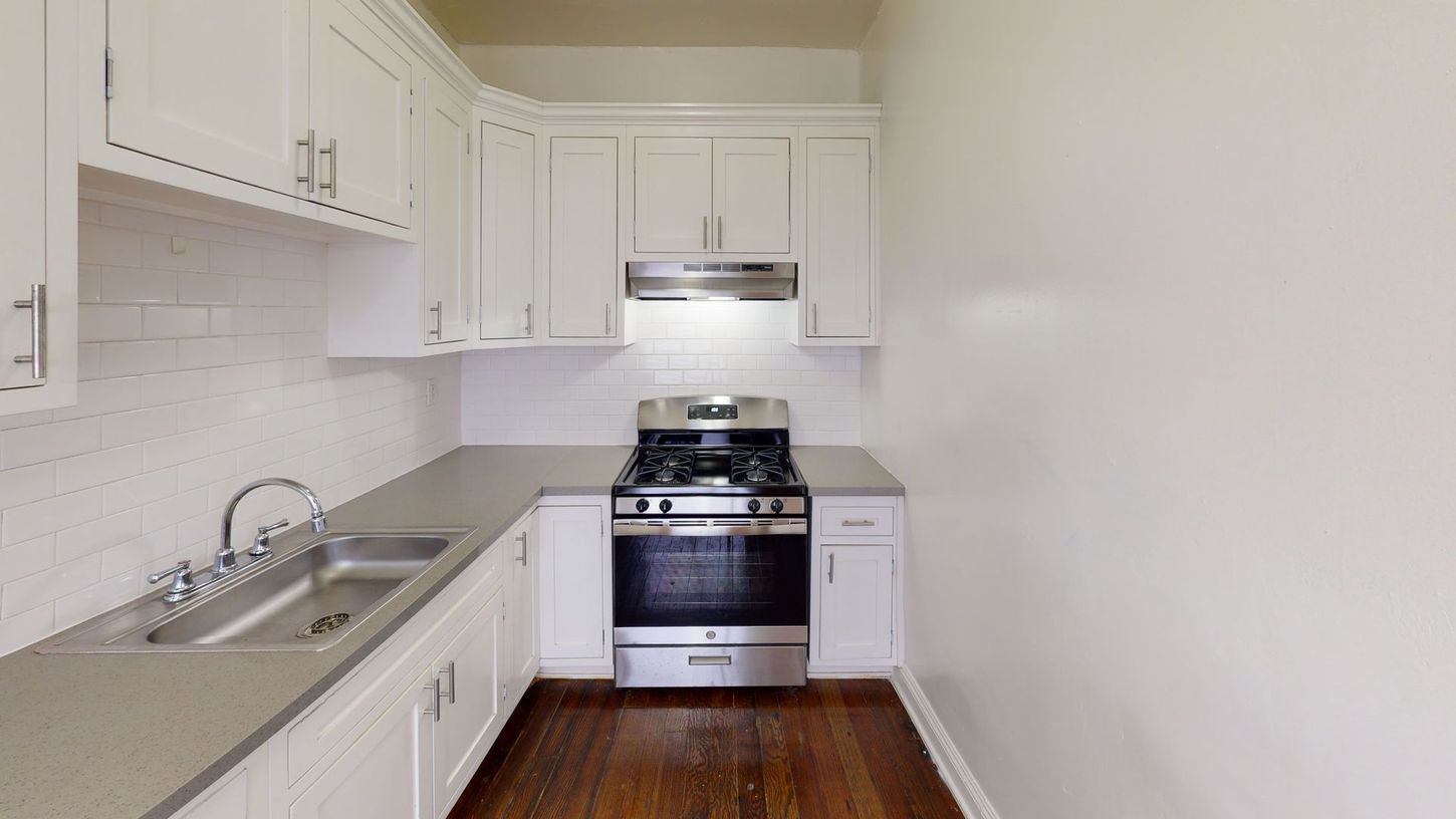 Small, white kitchen with stainless steel appliances and gray countertops; hardwood floor.