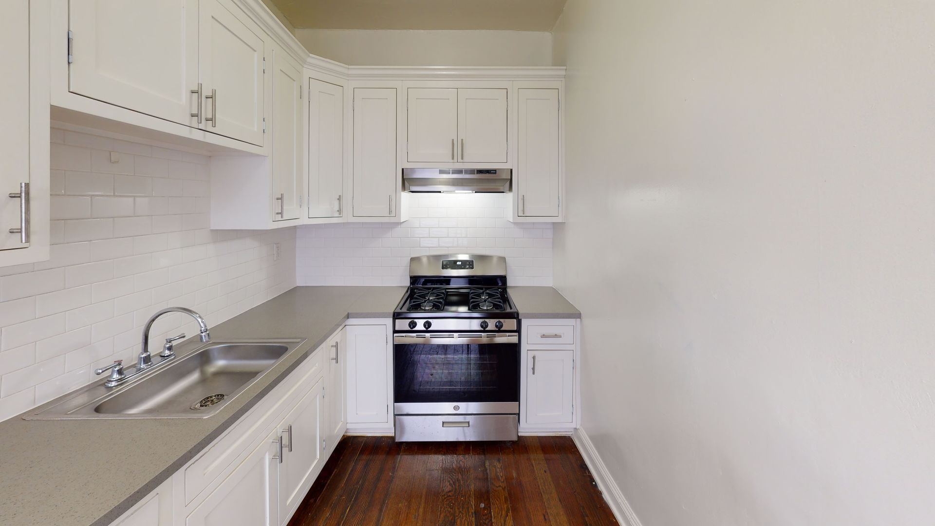Small, white kitchen with stainless steel appliances and gray countertops; hardwood floor.