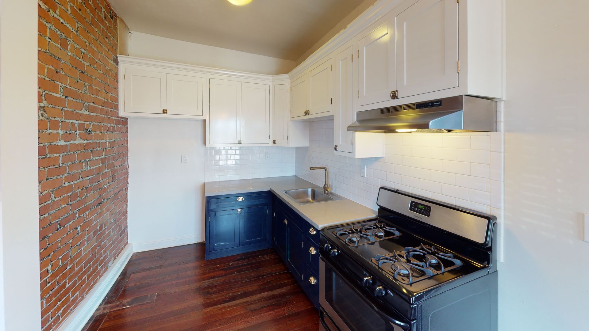 Small kitchen with blue and white cabinets, exposed brick wall, gas stove, and stainless steel sink.