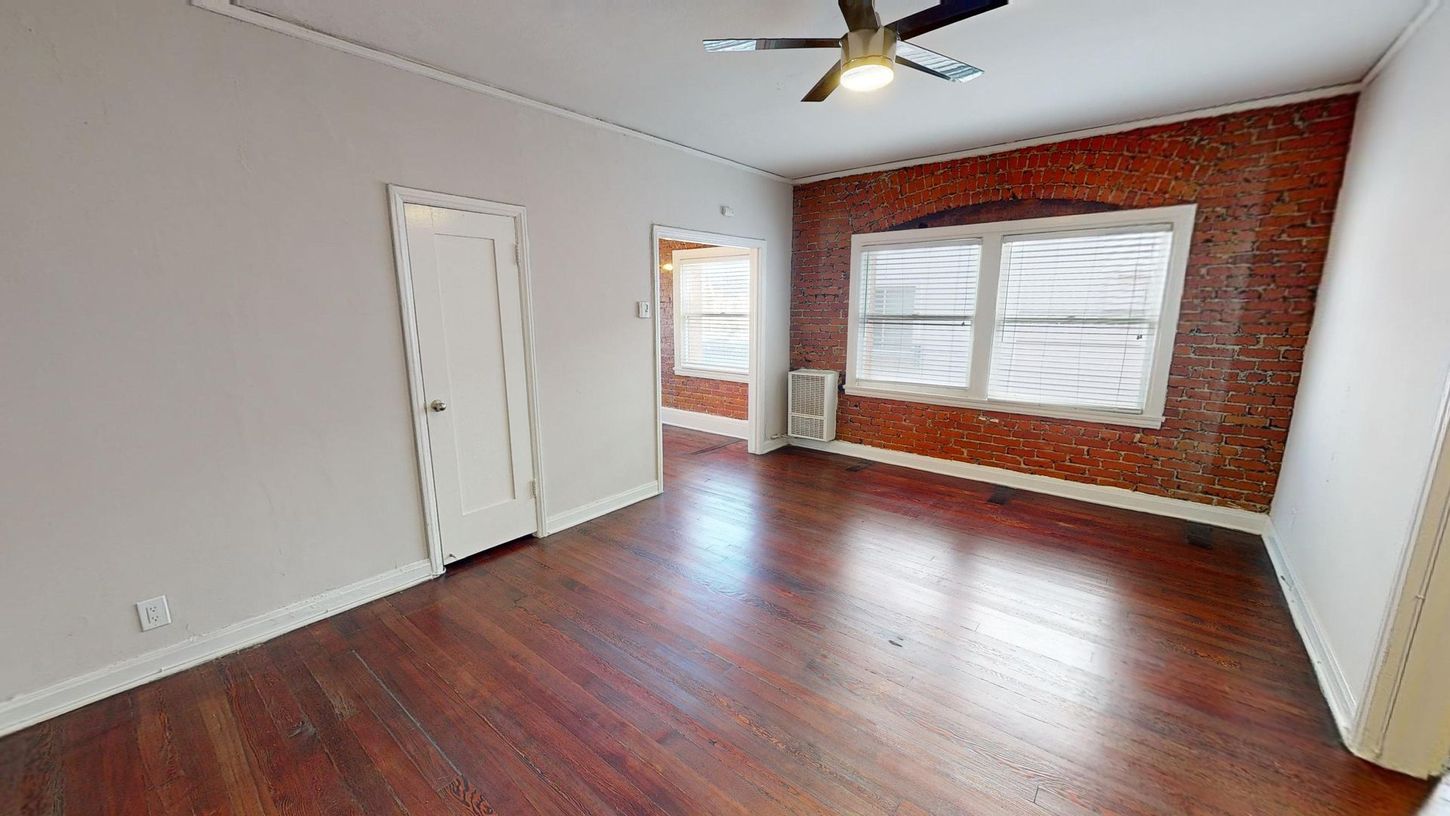 Interior of a room with hardwood floors, exposed brick wall, white door, and a ceiling fan.