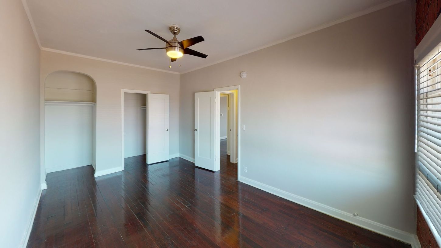 Empty bedroom with dark wood floors, light walls, ceiling fan, and open closet doors.