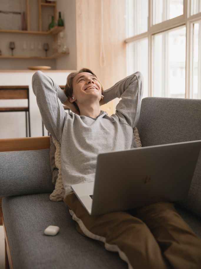 a man sits on a couch with his hands behind his head and a laptop