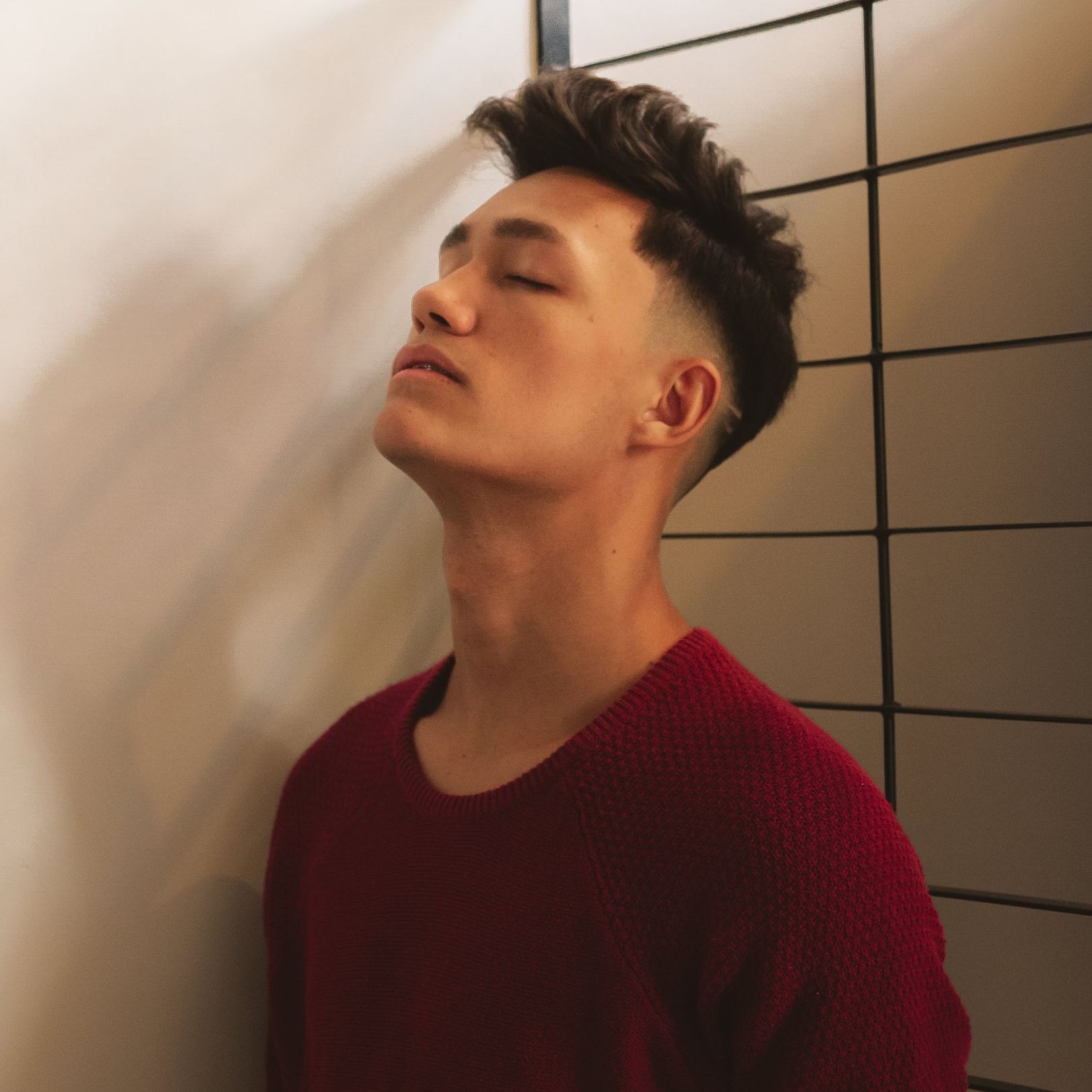 a young man in a red sweater leans against a wall with his eyes closed
