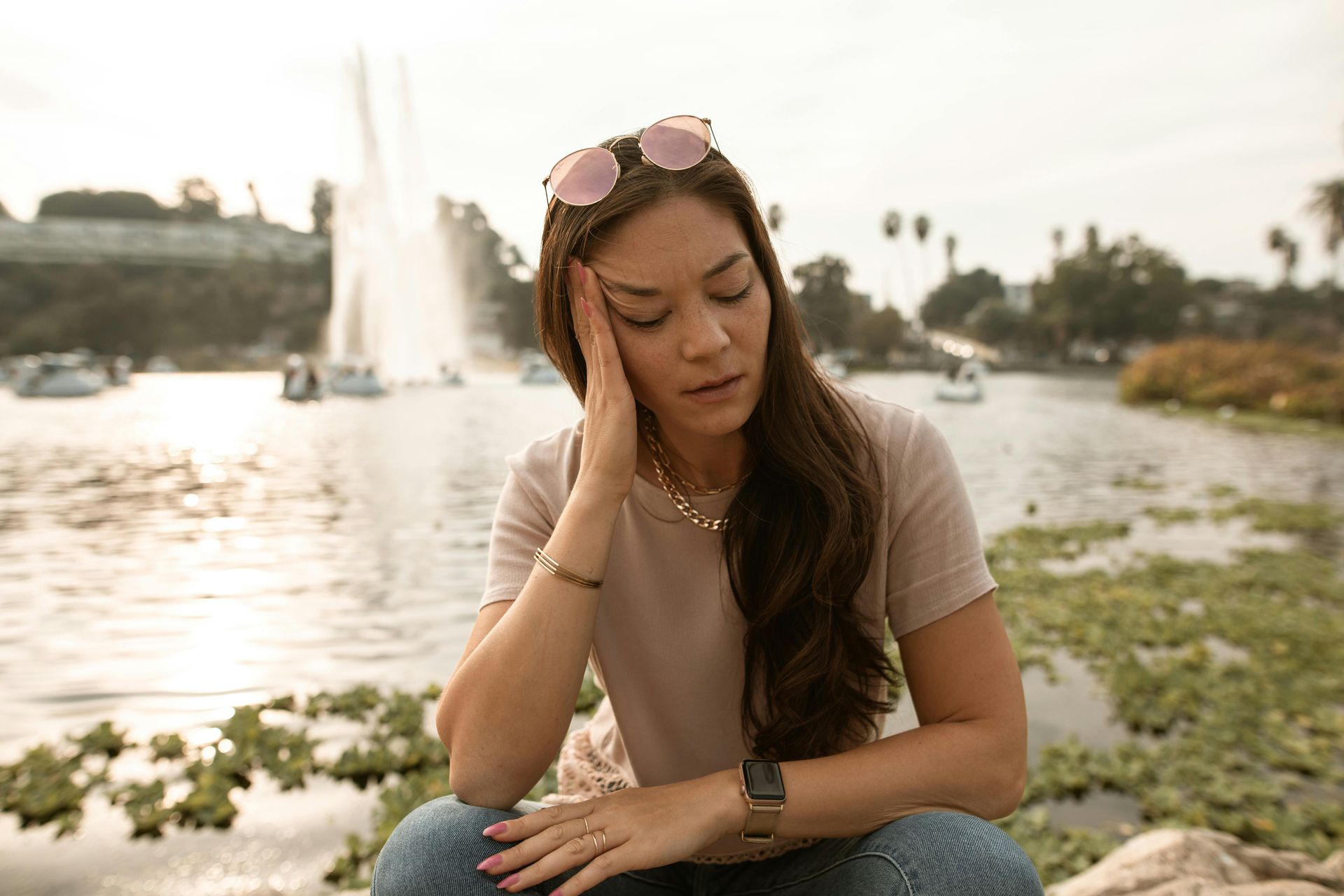 A woman is sitting in front of a lake with her hand on her forehead.