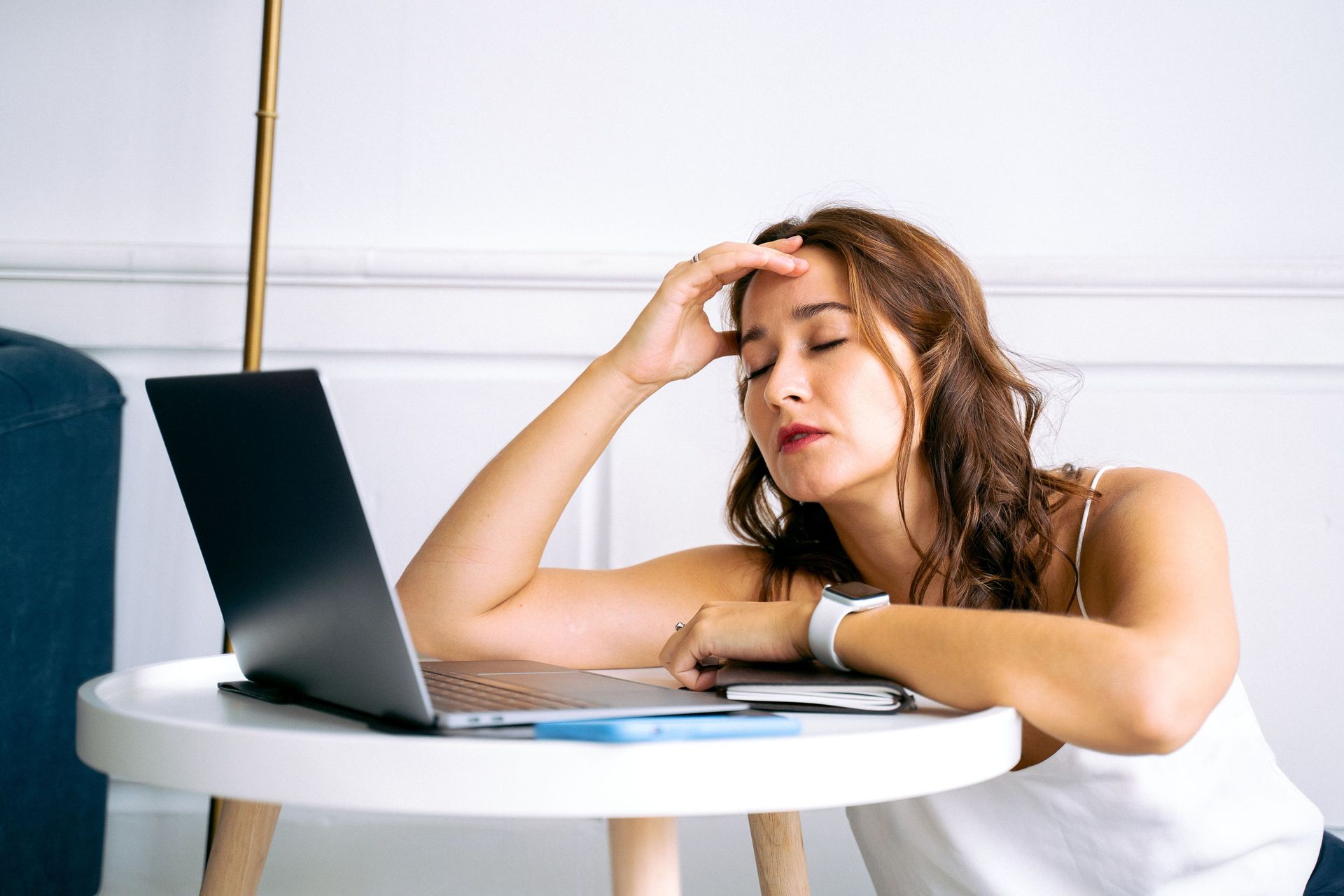 Woman at a table with a laptop, resting her head on her hand in a home office, looking tired and stressed.