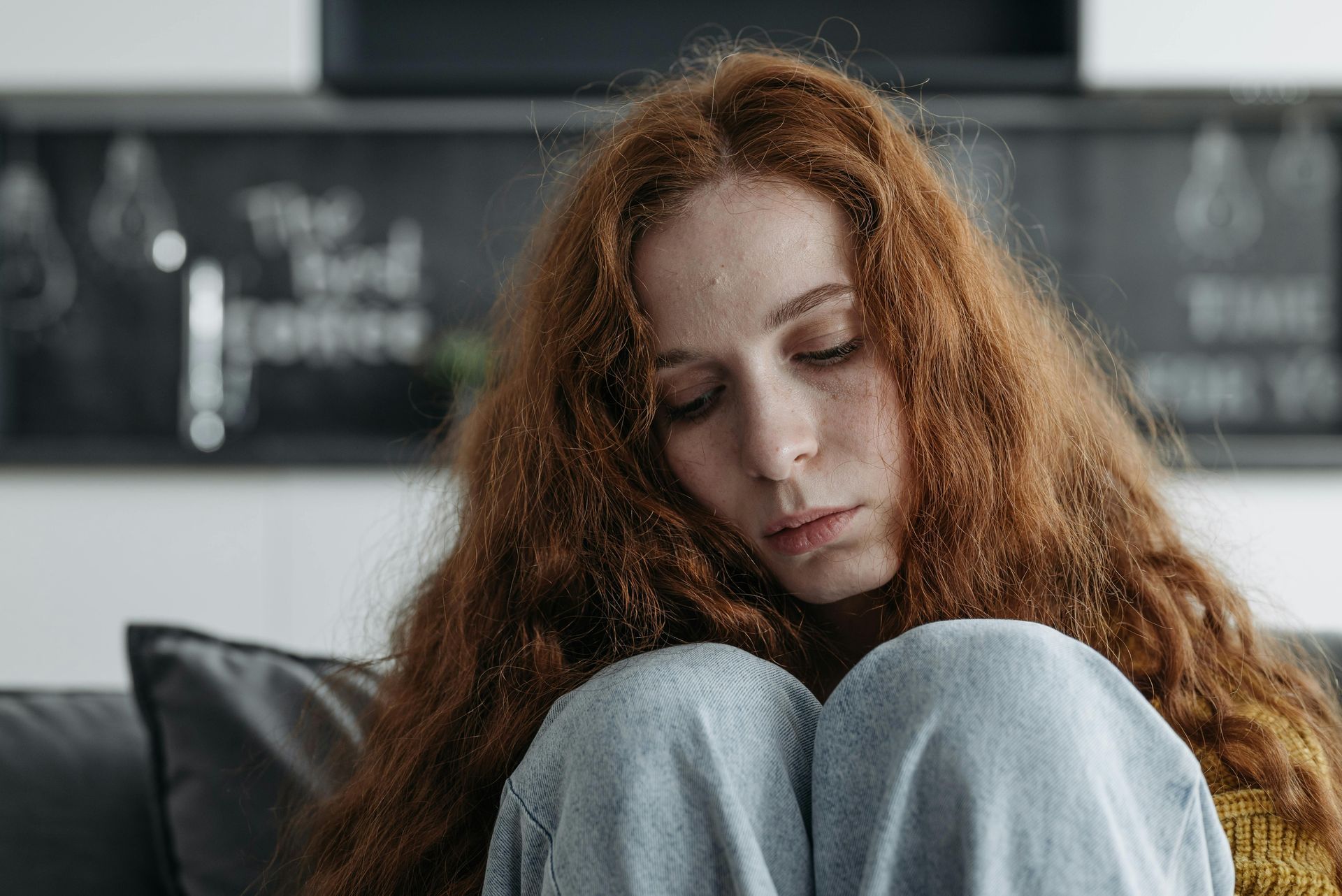 A person with long, curly auburn hair sits with their knees pulled up, looking down with a somber expression.