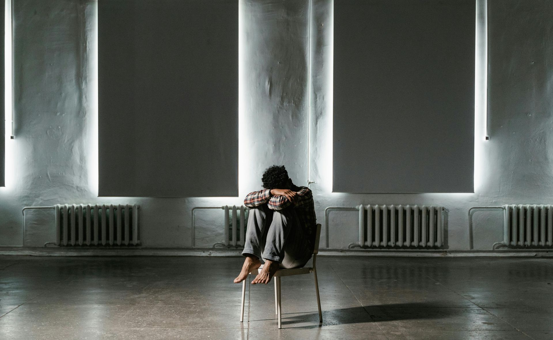 Lone person sitting on a reflective floor in a dim room with tall windows and radiators