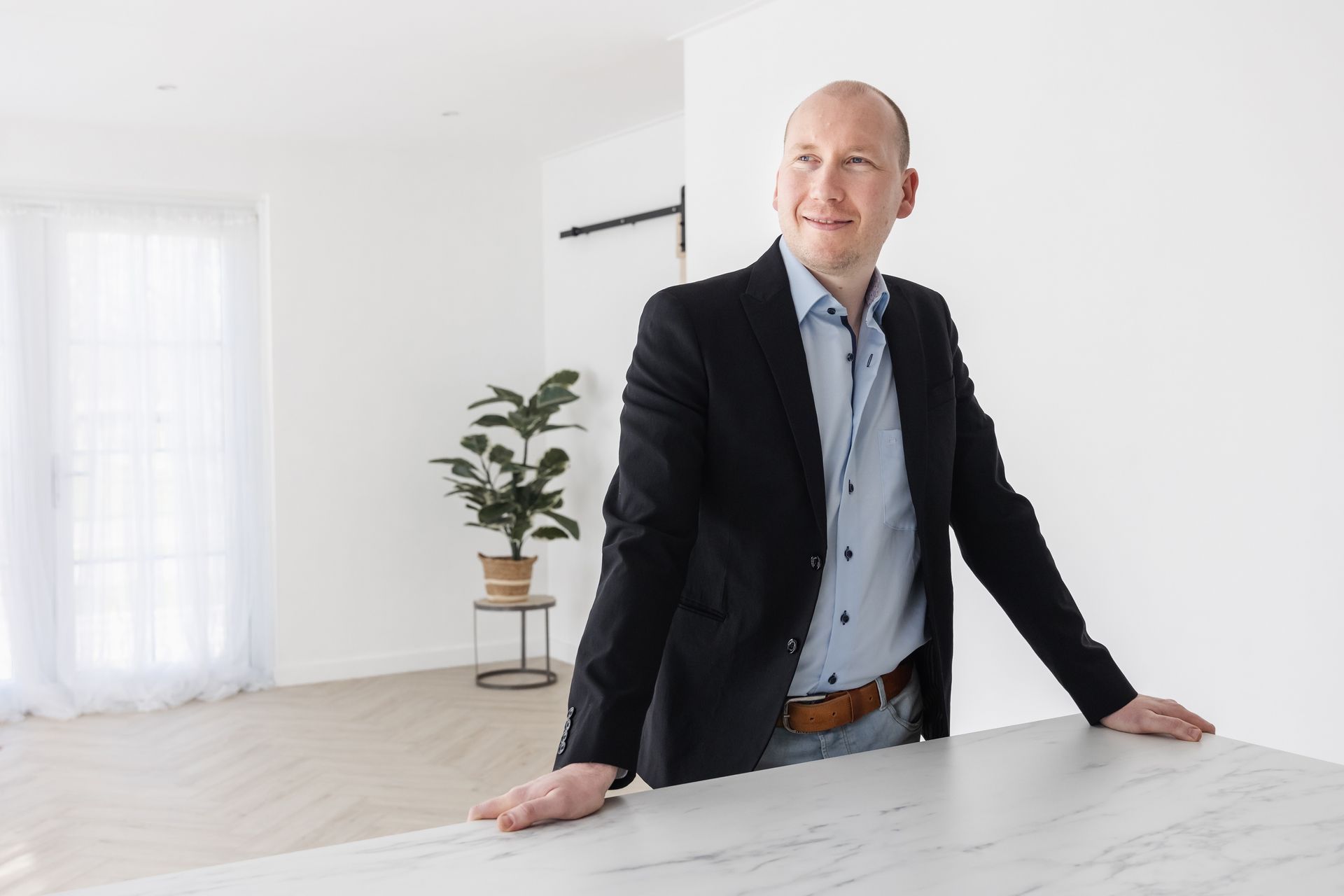 Een man in een blazer leunt glimlachend tegen een toonbank in een lichte, moderne ruimte.