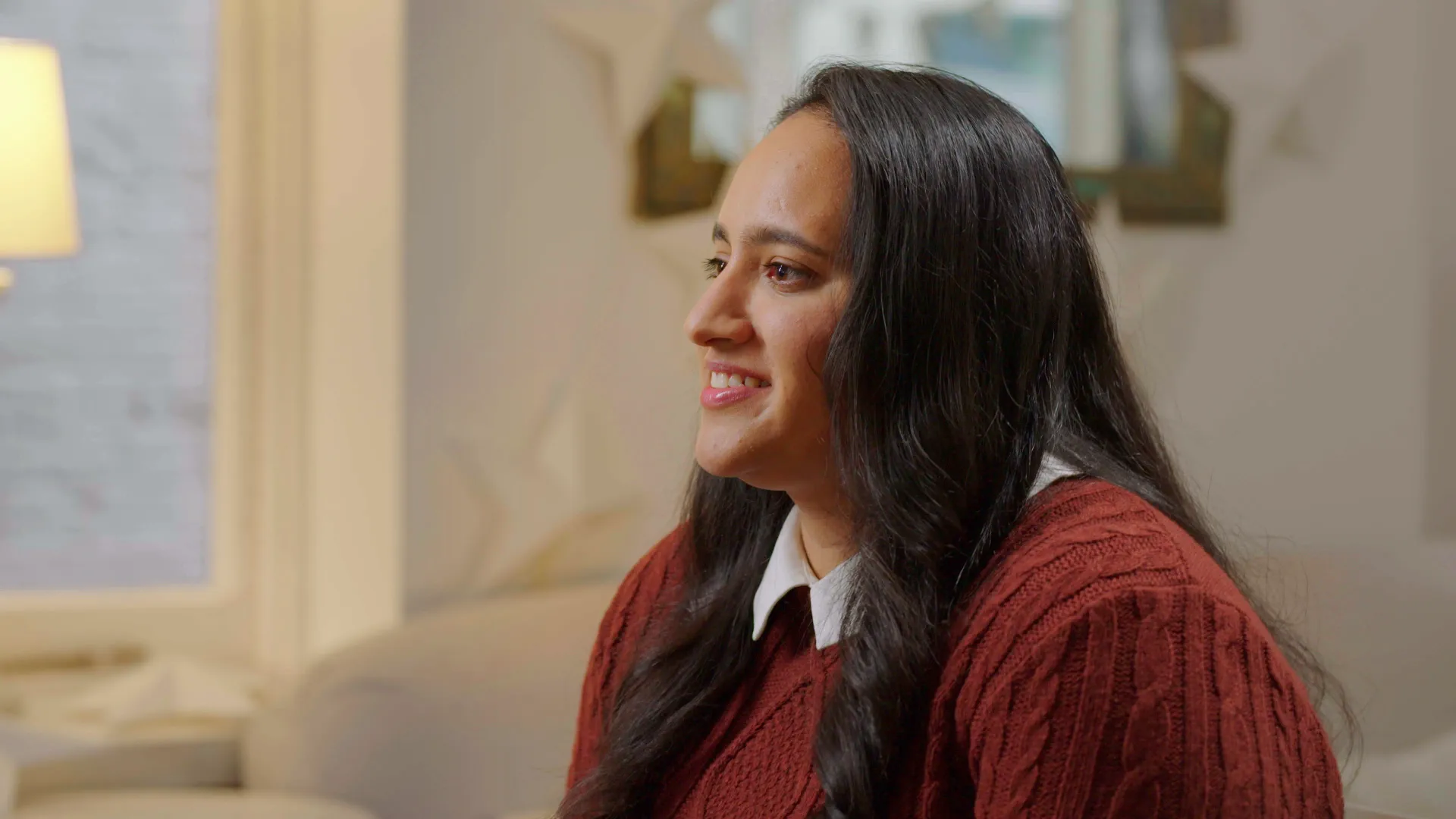 A woman in a red sweater is sitting on a couch and smiling.