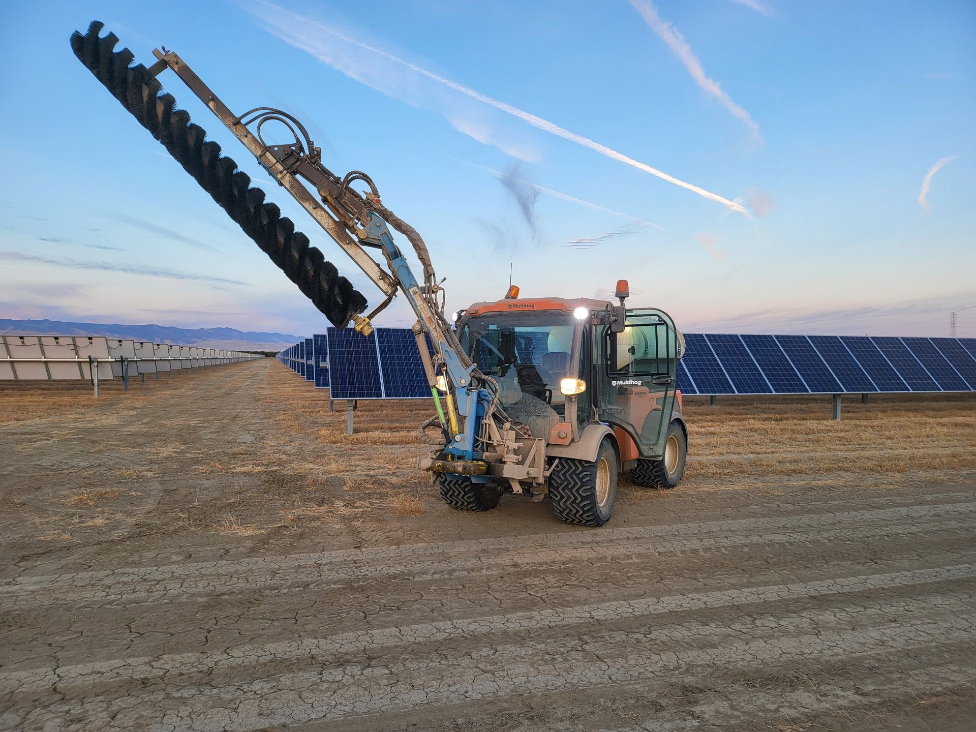 Tractor cleaning solar panels with an extending brush arm. Dusty field with panels and a blue sky in background.