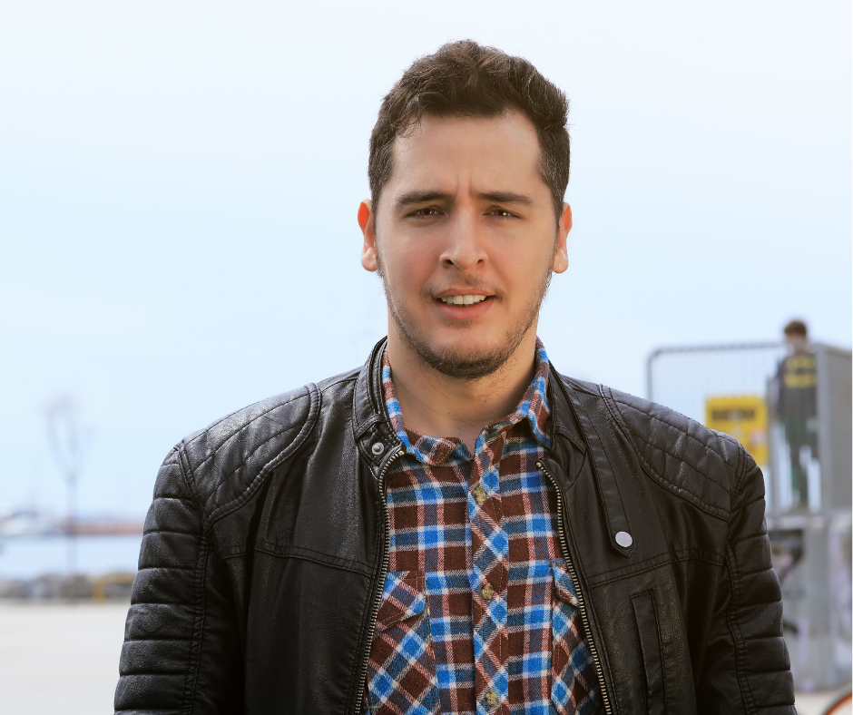 Man in leather jacket and plaid shirt, standing outdoors with a neutral expression.