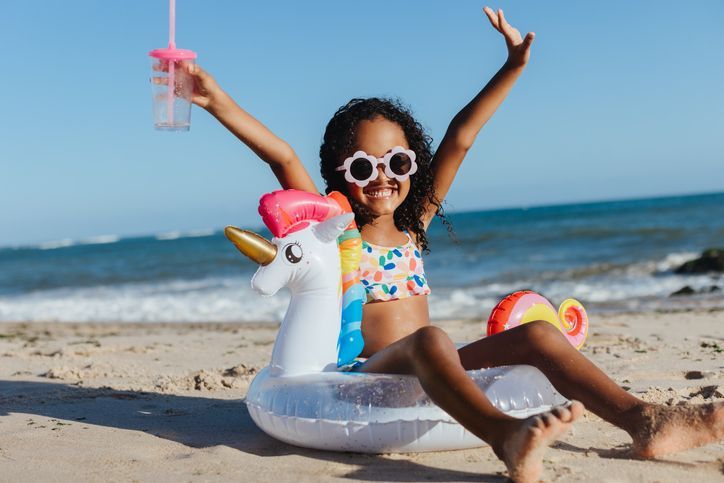 Girl in swimsuit on unicorn float at beach, arms raised, holding drink, smiling.
