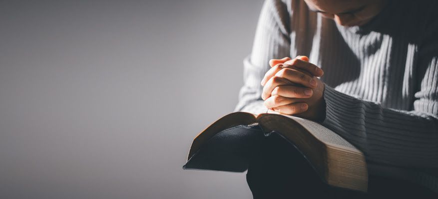 Person with hands clasped over an open Bible, head bowed in prayer.