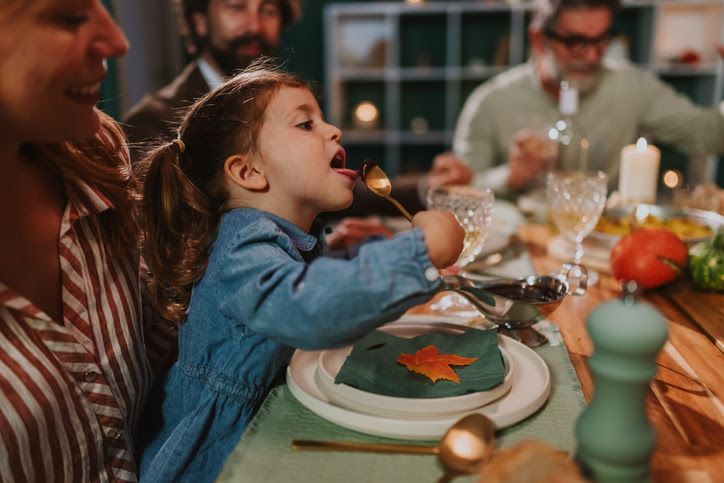 Girl at a dinner table tasting food from a spoon, surrounded by family, with a festive setting.