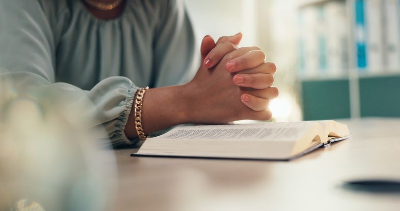 A lady is praying with her hands together on top of an open bible.