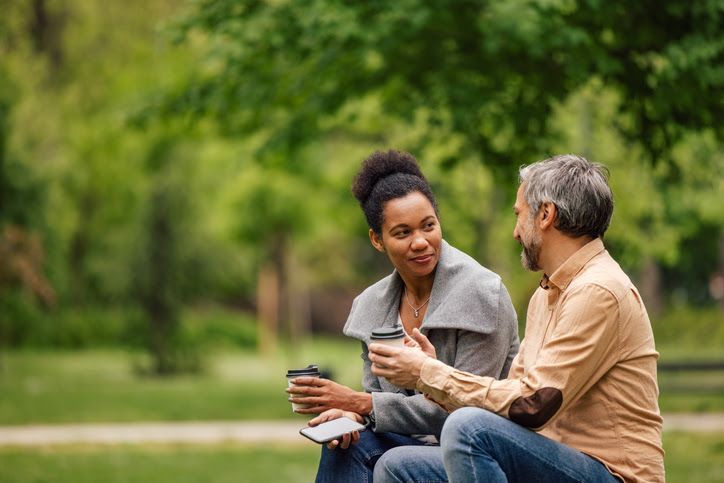 Woman and man in park, talking while holding coffee cups. Green trees in background.