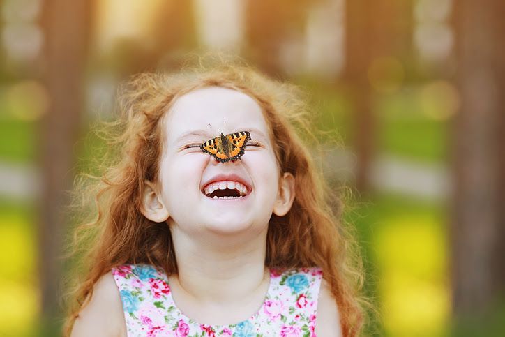 Girl with red hair smiles, a butterfly rests on her nose. Outdoors with soft focus background.