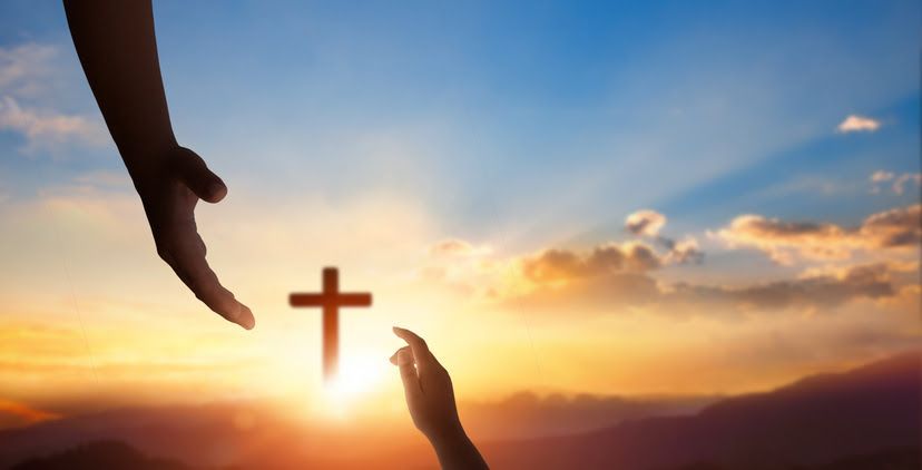 Hands reaching towards each other in front of a cross at sunset.