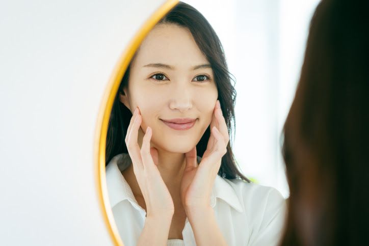 Woman smiles, touching her cheeks while looking in a mirror. She wears a white shirt in a bright room.