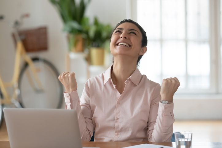Woman with arms raised in excitement, looking upward, next to a laptop at a desk.