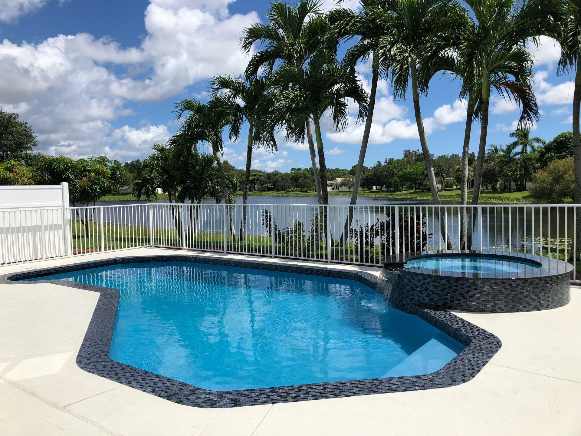 Blue pool and hot tub beside a white fence, with palm trees and a lake under a partly cloudy sky