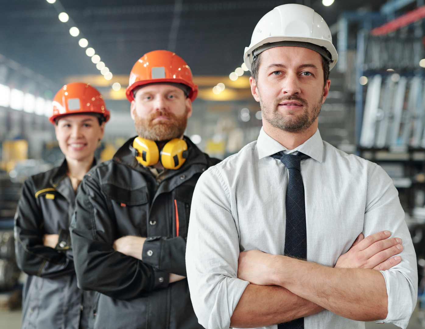 Three people in a factory. One in a white hard hat, dress shirt, and tie, arms crossed, smiling. Two in hard hats and workwear.