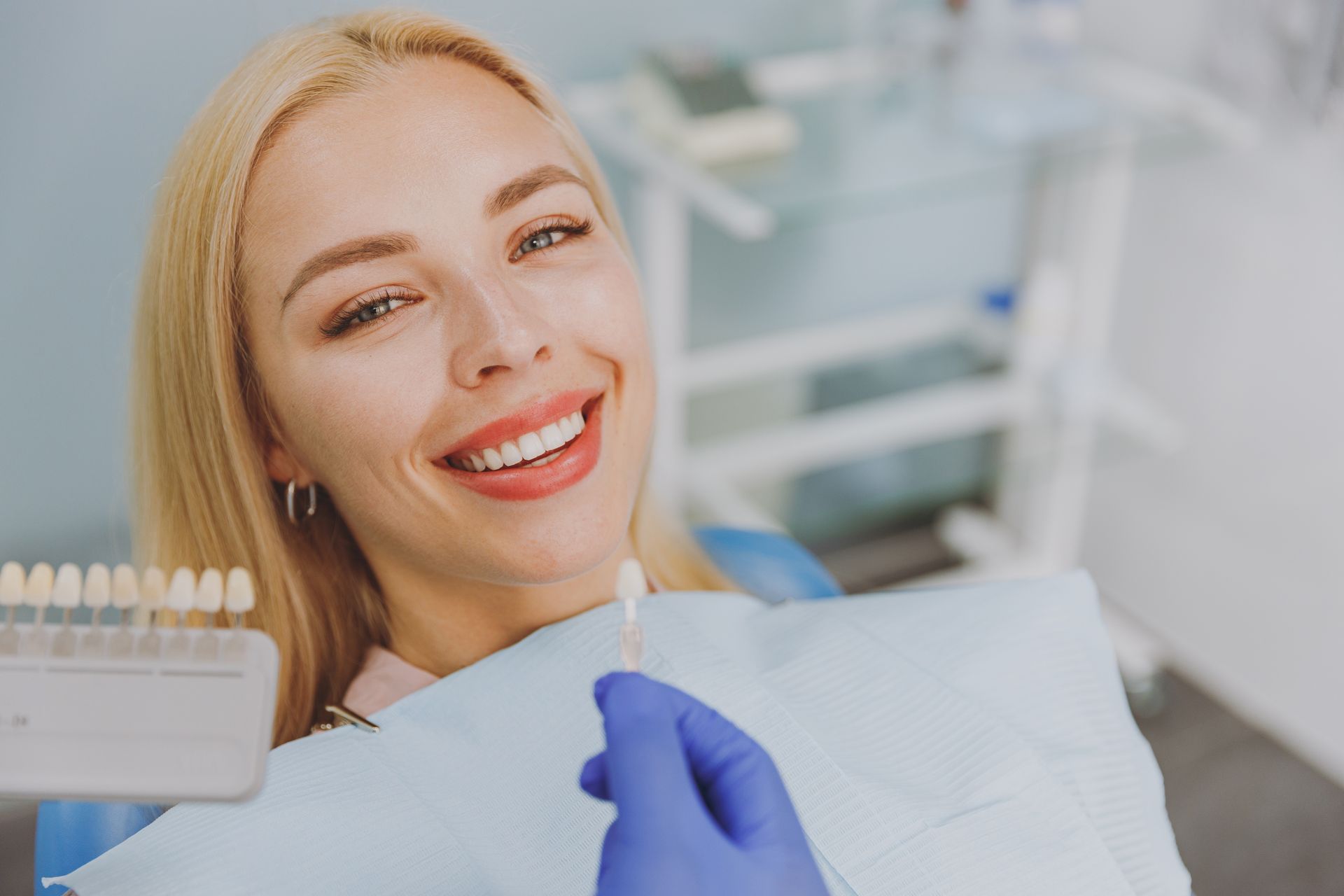 Woman smiling in dentist chair; dentist holding veneer sample.